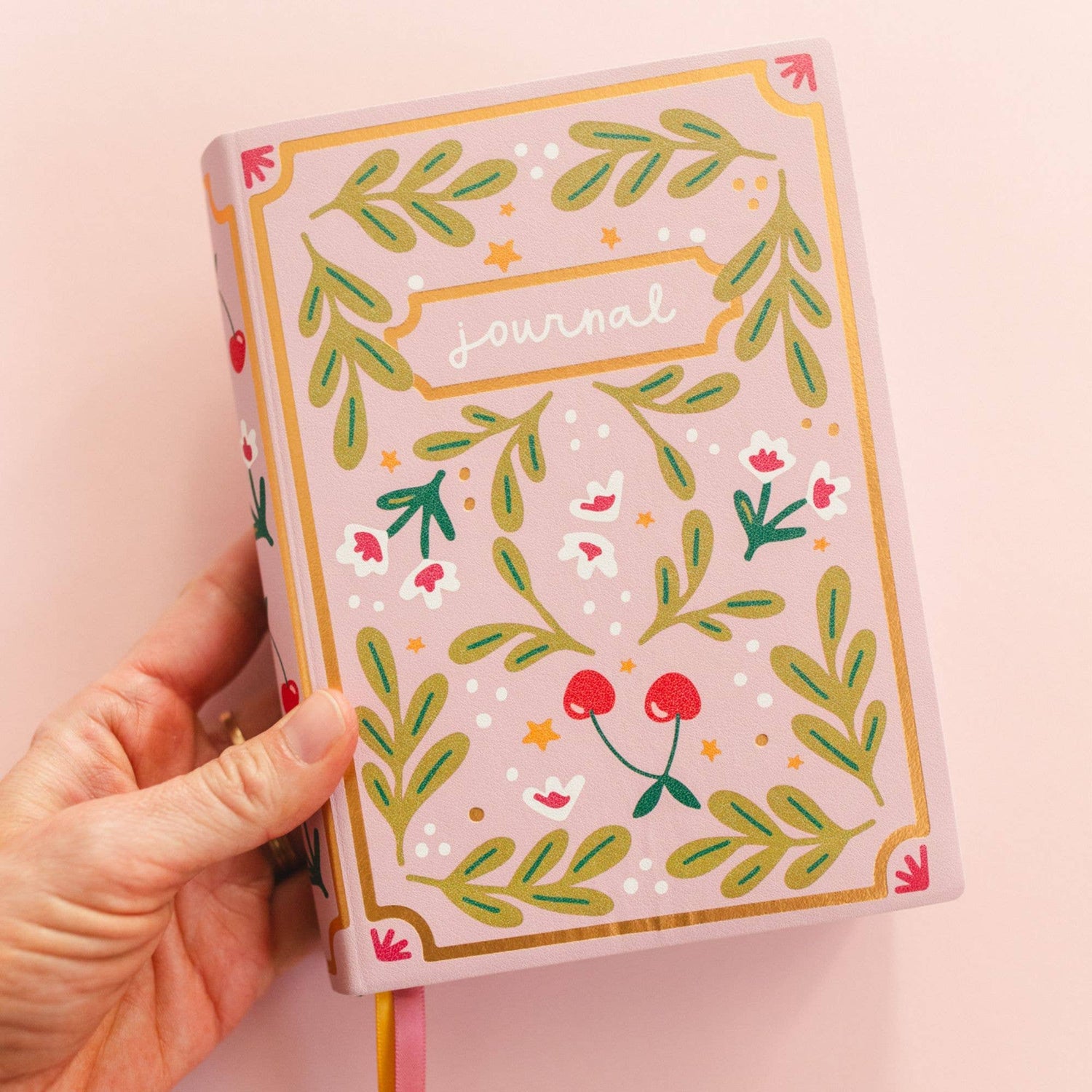 Hand holding a pink journal with floral designs on a light pink background