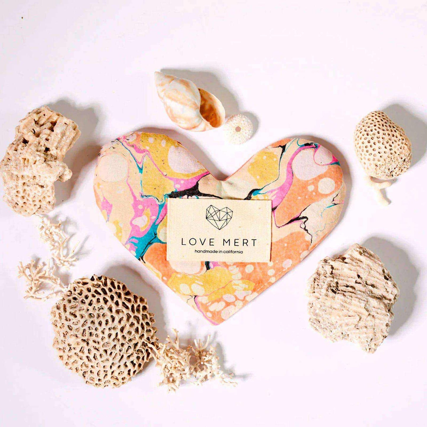 Heart-shaped Eye Love Pillow with marbled organic cotton and raw silk backing, surrounded by decorative shells and corals.