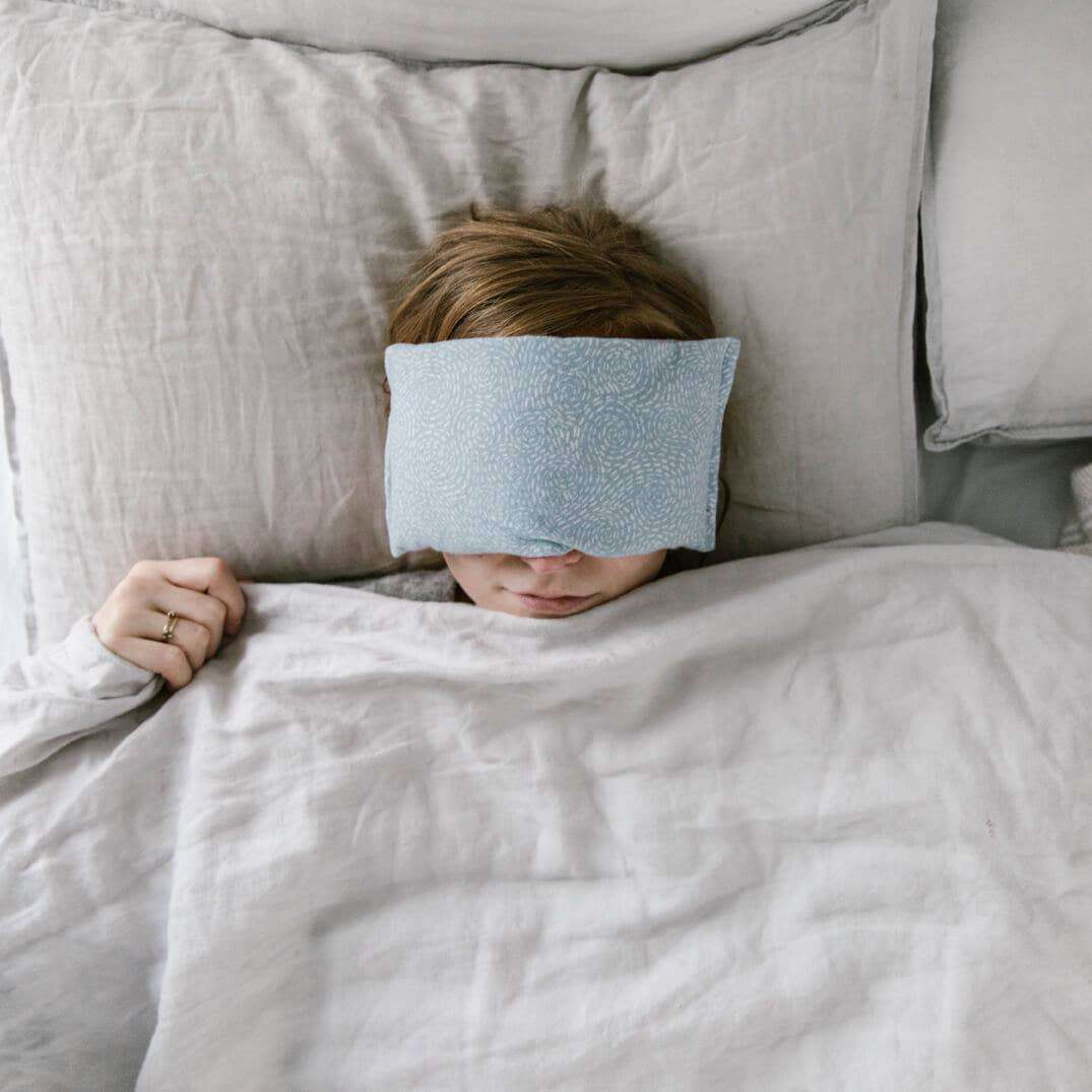 Lavender eye pillow on person relaxing in bed, promoting rest and aromatherapy benefits.