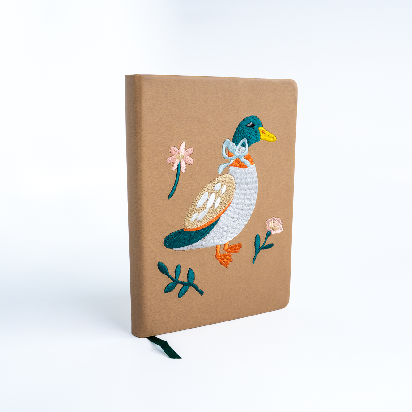A tan hardcover notebook with an embroidered mallard duck design on the front, featuring a green head, white body, pink bow, and orange beak, set against a clean white background.