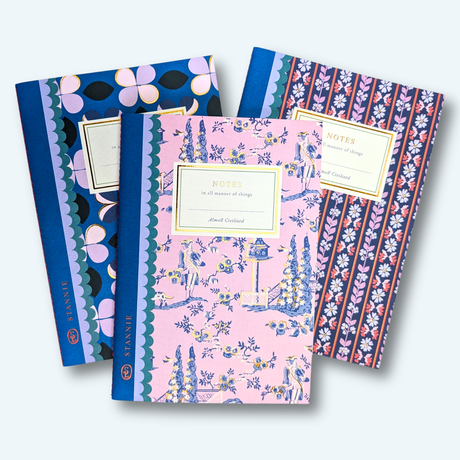 Three Anne Carroll Notes hardcover notebooks with floral and patterned covers, arranged in a fan shape against a light blue background.