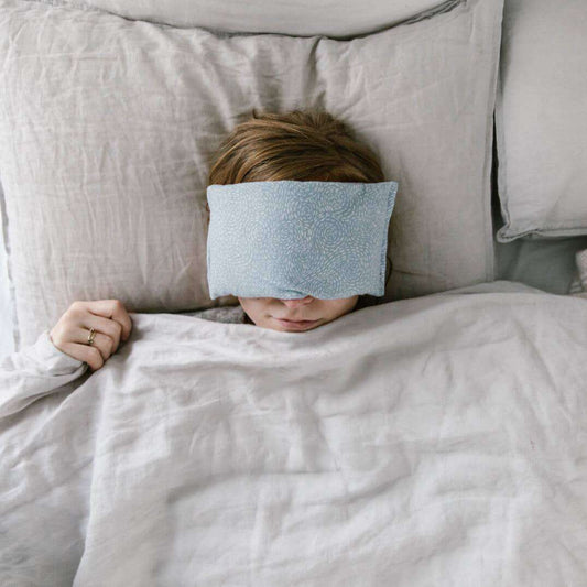 Lavender eye pillow on person relaxing in bed, promoting rest and aromatherapy benefits.