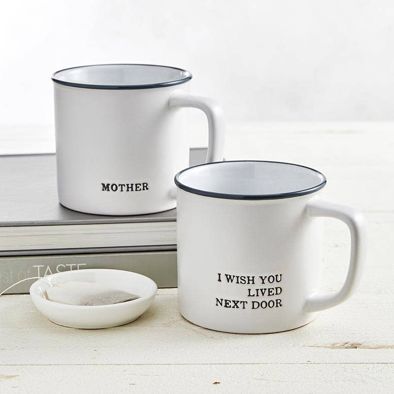 Two mugs on a table, one that says 'I Wish You Lived Next Door Mug' and another labeled 'MOTHER'.
