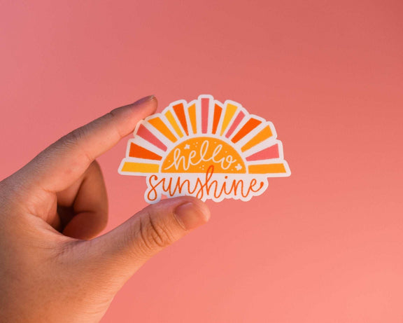 Hand holding a colorful Hello Sunshine Sticker with sunbeam design on a pink background.