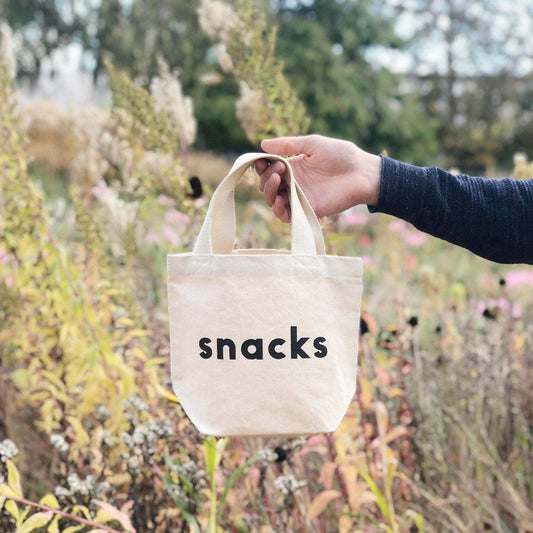 Natural canvas tote bag with "snacks" printed in black, ideal for kids' lunch and snacks, crafted in the UK.