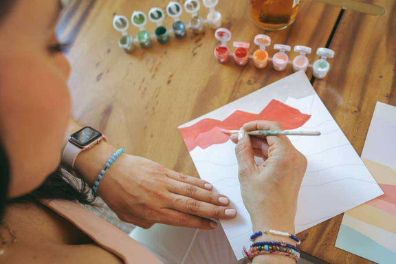 Person painting a red landscape with a Vibrant Butterflies Meditative Art Paint by Number Kit on a wooden table.