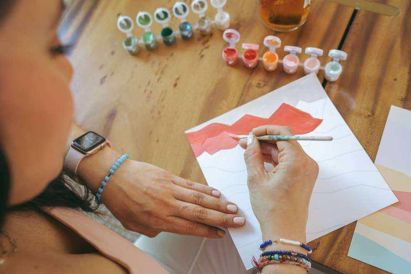 Person painting a red landscape with a Vibrant Butterflies Meditative Art Paint by Number Kit on a wooden table.